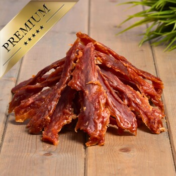 Ein Haufen hochwertiger Haustierleckereien aus getrockneten Fleischstreifen, auf einer Holzoberfläche mit einer grünen Pflanze im Hintergrund präsentiert.