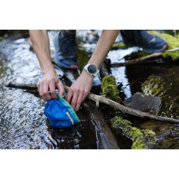 Eine Person hockt an einem Bach und hält eine blaue zusammenklappbare Haustierschüssel mit einem grünen Rand, umgeben von moosbedeckten Steinen und Ästen.