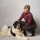 Eine Frau in einer kastanienbraunen Weste kniet neben einem großen, flauschigen schwarz-weißen Hund, beide schauen in die Kamera vor einem hellgrauen Hintergrund.