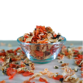 Eine klare Glasschüssel hält bunte getrocknete Gemüseflocken in Orange, Gelb und Grün, mit verstreuten Flocken auf einer hellen Oberfläche.