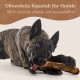 Ein gestromter Hund kaut auf einem Olivenholzstock, mit dem Text darüber: "Olivenholz-Kaustab für Hunde" und "100% natürlich, chemie- und zusatzstofffrei."