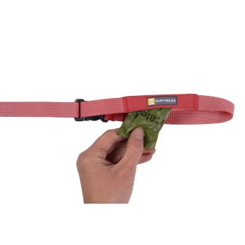 Eine Hand zieht einen grünen Müllbeutel von einer roten Hundeleine mit einem Ruffwear-Logo, entworfen für eine einfache Müllentsorgung während der Spaziergänge.