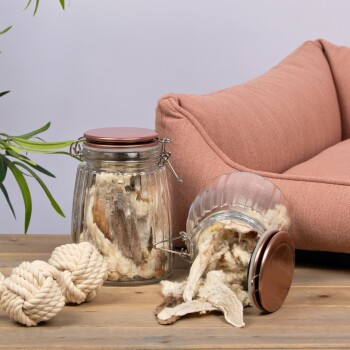 Zwei Glasbehälter mit Hundeleckerlis stehen auf einem Holztisch neben einem rosa Haustierbett und einem Paar Seilspielzeuge.