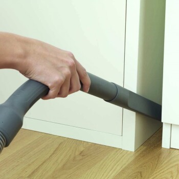 Eine Hand hält einen Staubsaugerschlauch, der unter einem Schrank auf einem Holzboden greift, mit Fokus auf das Reinigen enger Räume.