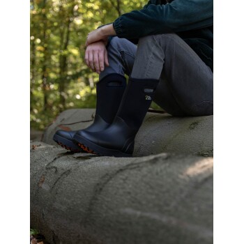 Eine Person sitzt auf einem Baumstamm in einem Wald, trägt schwarze Gummistiefel mit orangefarbenen Sohlen und graue Hosen und ruht ihre Hände auf den Knien.