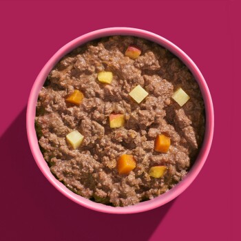 Eine rosa Schüssel, gefüllt mit Haustierfutter, das eine klumpige, braune Mischung mit kleinen Stücken orangefarbener und gelber Gemüse enthält.
