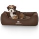 Ein kleiner, flauschiger Welpe sitzt bequem in einem braunen Hundebett, auf dem das Logo "KNUFFELWUFF" deutlich sichtbar ist.