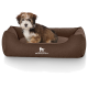 Ein kleiner, flauschiger Welpe sitzt bequem in einem braunen Hundebett, auf dem das Logo "KNUFFELWUFF" deutlich sichtbar ist.