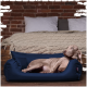 Ein entspannter Hund liegt in einem gemütlichen navyblauen Haustierbett, vor einer rustikalen Ziegelwand und einem ordentlich gemachten Bett mit einer gestrickten Decke.