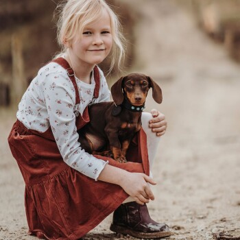 Ein Kind in einem blumigen Langarmhemd und einem rostfarbenen Kleid sitzt auf dem Boden und hält einen kleinen braunen Dackel mit einem blauen Halsband.