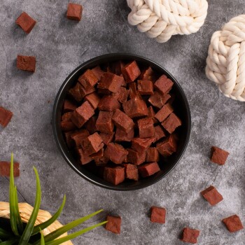 Eine schwarze Schüssel gefüllt mit kleinen, gewürfelten Hundeleckerlis, umgeben von verstreuten Stücken, einer grünen Pflanze und zwei geknoteten Seilspielzeugen auf einer grauen Oberfläche.