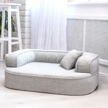 Ein schwarz-brauner Dackel ruht auf einem gemütlichen grauen Hundebett mit zwei kleinen Kissen, in einem hellen, luftigen Raum mit weißen Vorhängen.