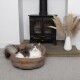 Eine flauschige grau-weiße Katze liegt in einem gemütlichen braunen Haustierbett mit der Aufschrift "Scruff's", das sich in der Nähe eines schwarzen Holzofens und einer dekorativen Vase befindet.