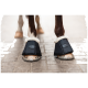 Nahaufnahme der Hufe eines Pferdes, das schwarze schützende Hufschuhe mit weißem Fleecefutter trägt und auf einer Ziegeloberfläche steht.