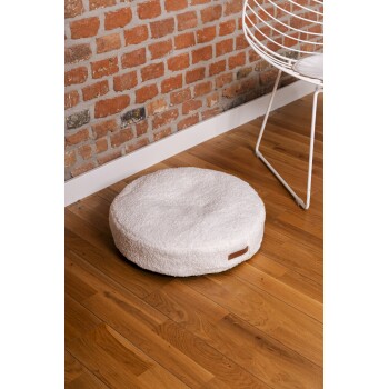 Ein rundes, flauschiges weißes Haustierbett steht auf einem Holzboden, neben einem modernen weißen Drahtstuhl und einer Ziegelwand.