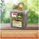 Transparenter Behälter mit der Aufschrift 'PANTO Winterbox' mit Vogelfutter, zwei Netzfettkugeln und einem Samenring auf einem Tisch, grüner Bokeh-Hintergrund