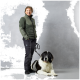 Eine Person in einer grünen Jacke und schwarzen Hosen steht neben einem großen, flauschigen schwarz-weißen Hund an der Leine, vor einem grauen Hintergrund.