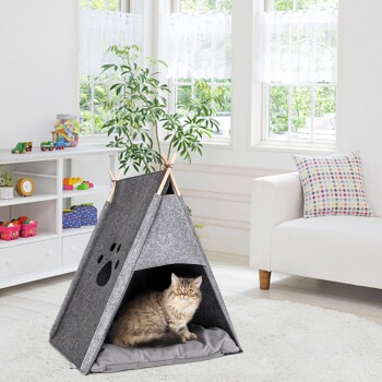 Eine flauschige Katze sitzt in einem grauen Filz-Haustier-Tipi mit einem Pfotenabdruckdesign, in einem hellen Raum mit großen Fenstern und einer gemütlichen Atmosphäre.