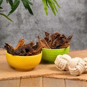 Zwei Schalen mit Hundeleckerlis: eine gelbe Schale mit dunklen knusprigen Stücken und eine grüne Schale mit helleren, kaubaren Stückchen, neben einem geknoteten Seilspielzeug.