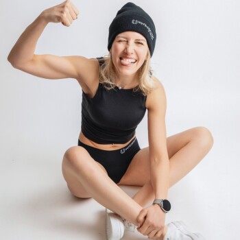 Eine Person in einem schwarzen Crop-Top und Unterwäsche beugt ihren Arm auf dem Boden, trägt eine mit Logo versehene schwarze Mütze.