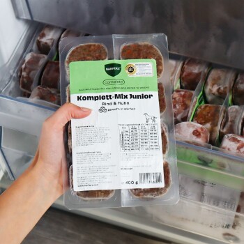 Hand hält gefrorene BARFER'S ‚Komplett‑Mix Junior Rind & Huhn‘ Hundefutter‑Packung (400 g) in einer Gefrierschublade.