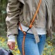 Torso einer Person in beige Jacke und Jeans mit an eine Gürtelschlaufe befestigter, leuchtend orangefarbener Hundeleine, unscharfer Waldhintergrund.