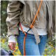 Torso einer Person in beige Jacke und Jeans mit an eine Gürtelschlaufe befestigter, leuchtend orangefarbener Hundeleine, unscharfer Waldhintergrund.