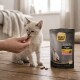 Kitten being hand-fed treats beside a bag labeled "SELECT GOLD Snacks Pure Huhn Kitten" on a wooden floor.