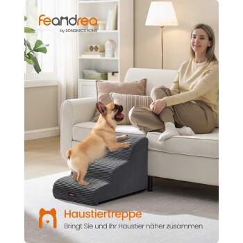 Kleiner hellbrauner Hund klettert graue Haustiertreppe neben einer Frau, die auf einem weißen Sofa in einem hellen Wohnzimmer sitzt.