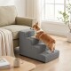 Braun-weißer Corgi klettert eine graue Haustiertreppe mit drei Stufen neben einem beigen Sofa in einem hellen Wohnzimmer.