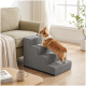 Braun-weißer Corgi klettert eine graue Haustiertreppe mit drei Stufen neben einem beigen Sofa in einem hellen Wohnzimmer.