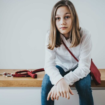 Eine Person sitzt auf einer Holzbank, trägt ein weißes Hemd und blaue Jeans, mit einer roten Tasche und einer roten Hundeleine neben sich.