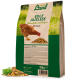 Braune Henne, die neben einem Haufen Hühnerfutter auf einem Sack mit der Aufschrift "Hühner Land Edle Auslese Hühnerfutter mit Oregano," 10kg steht.