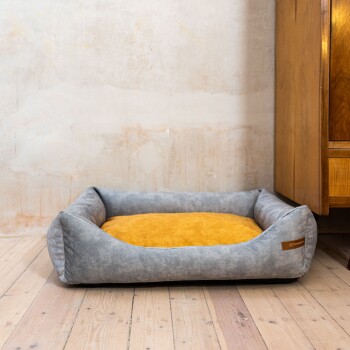 Ein gemütliches Haustierbett mit einem grauen Stoffbezug und einem weichen gelben Kissen, auf Holzboden neben einem Holzschrank platziert.