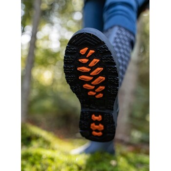 Nahaufnahme der Sohle eines schwarzen Gummistiefels mit orangefarbenen Traktionsmustern, vor einem verschwommenen Waldhintergrund.