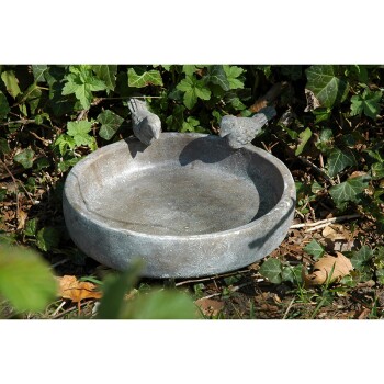 Ein dekorativer Steinvogelbad mit zwei kleinen Vogelskulpturen, die auf dem Rand sitzen, umgeben von grünem Laub.