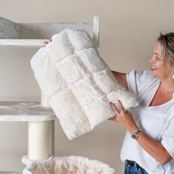 Eine Frau hält ein plüschiges, cremefarbenes Haustierbettkissen mit einem gesteppten Design und steht neben einem Katzenbaum.