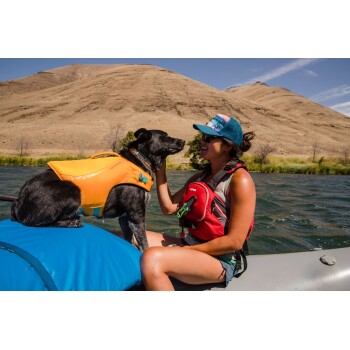 Ein schwarzer Hund in einer orangefarbenen Schwimmweste sitzt auf einem blauen Floß, während eine Frau in einer roten Weste mit ihm am Fluss und den Hügeln interagiert.