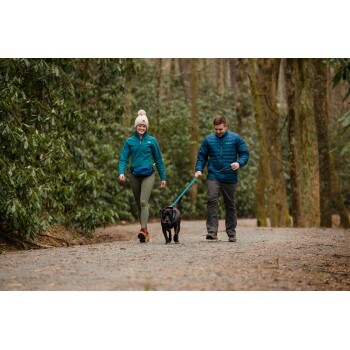 Ein Mann und eine Frau gehen mit einem schwarzen Hund auf einem Kiesweg, umgeben von Bäumen und Grün, beide in Outdoor-Bekleidung gekleidet.