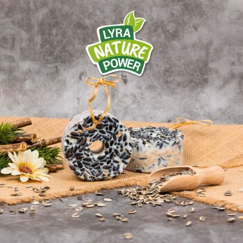 Zwei runde Vogelfutterkuchen auf Jute, einer mit einem Loch, umgeben von Sonnenblumenkernen und einem Holzlöffel. Text: "LYRA NATURE POWER."