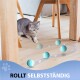 Eine verspielte graue Katze hockt auf einem Holzboden und beobachtet rollende blaue Bälle mit "ROLLT SELBSTÄNDIG" am Boden.