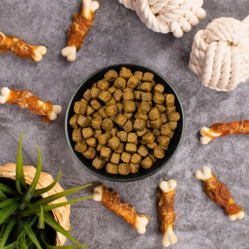 Eine schwarze Schüssel, gefüllt mit kleinen, grünlich-braunen Haustierfutter-Kroketten, umgeben von Hundekau-Knochen und einem geflochtenen Pflanzenkübel mit Grünpflanzen.