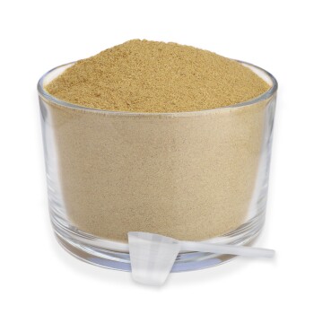 Ein klarer Glasbehälter, gefüllt mit hellbraunem Pulver, begleitet von einem kleinen weißen Messlöffel, der daneben liegt.