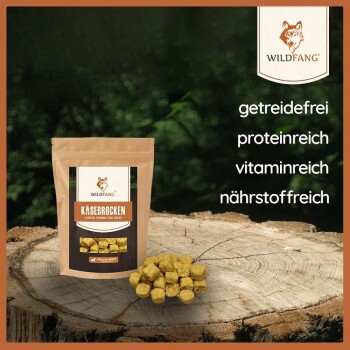 Eine Tüte Wildfang Käsebrocken für Hunde liegt auf Holz, mit verstreuten gelben Käsestücken und Text: "getreidefrei," "proteinreich," "vitaminreich."