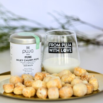 Ein Behälter mit "Puja Dog Pure Milky Churpi Pops" neben einem Glas Milch und einem Teller mit runden Hundeleckerlis. Das Glas liest "VON PUJA MIT LIEBE."