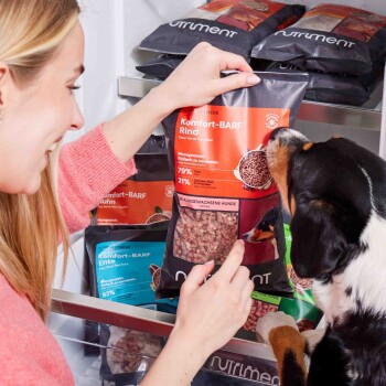 Eine Person hält einen "Komfort-BARF Rind" Hundefutterbeutel mit rotem Etikett. Ein Hund schnüffelt daran, mit anderen Hundefutterbeuteln im Hintergrund.