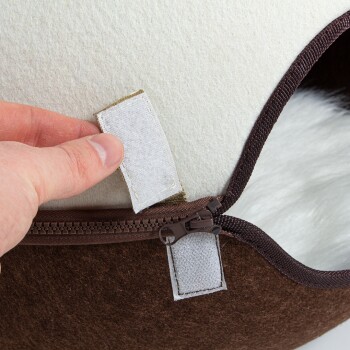 Die Hand einer Person hält ein Stoffetikett in der Nähe eines Reißverschlusses auf einem Haustierbett, das das braun-creamfarbene Farbschema und das weiche Innere zeigt.