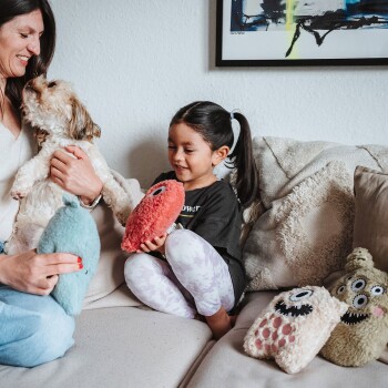 Eine Frau hält einen kleinen Hund, während sie auf einer Couch sitzt, neben einem Kind, das ein Plüschtier hält. Bunte Plüschtiere sind auf der Couch verstreut.