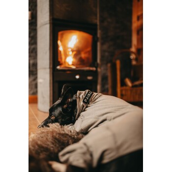 Ein schwarzer Hund, der einen grauen Pullover mit dem Etikett "PAIKKA" trägt, liegt bequem auf einer braunen, pelzigen Decke vor einem leuchtenden Kamin.