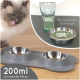 Eine flauschige graue Katze leckt sich die Lippen neben einem grauen doppelten Fressnapfset mit zwei Edelstahlschalen und einem "100ML" Glas.